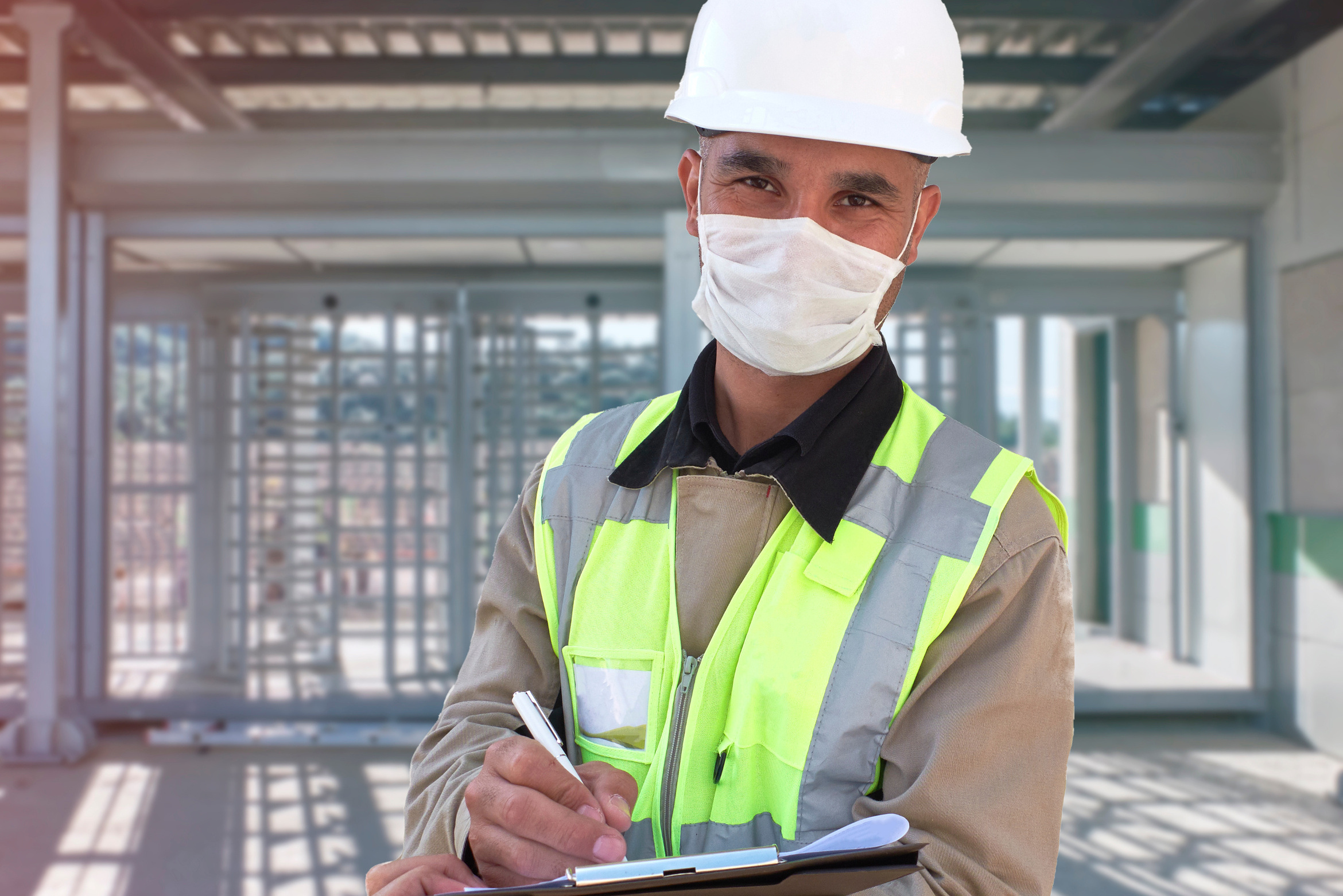 Bauarbeiter mit Schutzmaske, Sicherheitsweste und Helm steht auf einer Baustelle und schreibt Notizen auf ein Klemmbrett.